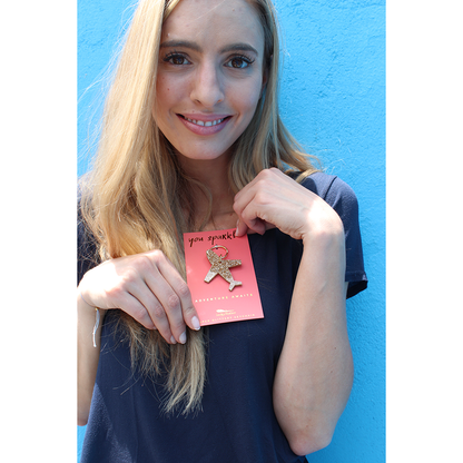 A smiling woman holds a keychain with a glittery airplane charm attached to a card that reads 'You Sparkle' and 'Adventure Awaits.'