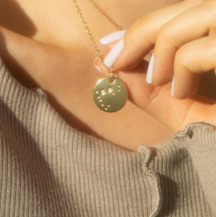 A person with white painted fingernails holds a gold constellation pendant on a gold chain.