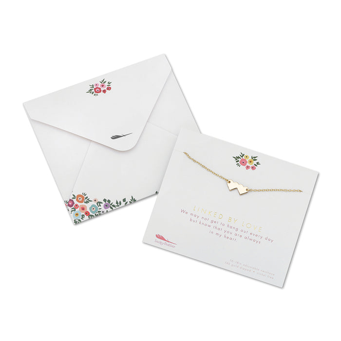 An envelope with floral decoration and a 'Linked by Love' card displaying a gold heart necklace are on a white background.