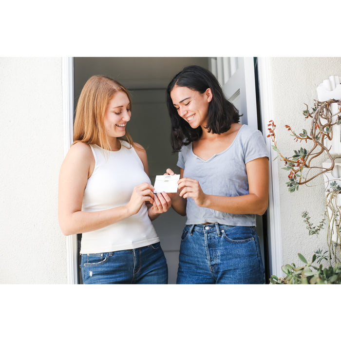 Two smiling women stand in a doorway looking at a small card with a necklace.