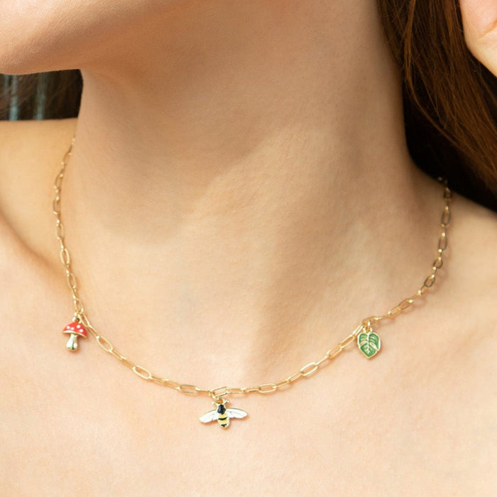 A person wears a gold chain necklace with mushroom, bee, and leaf charms.