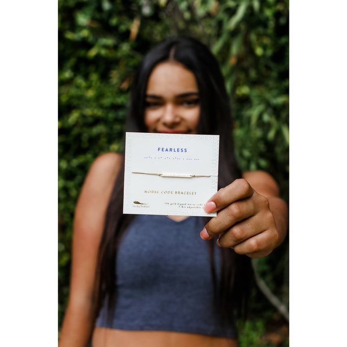 A person holds up a card displaying a 'Fearless' morse code bracelet.