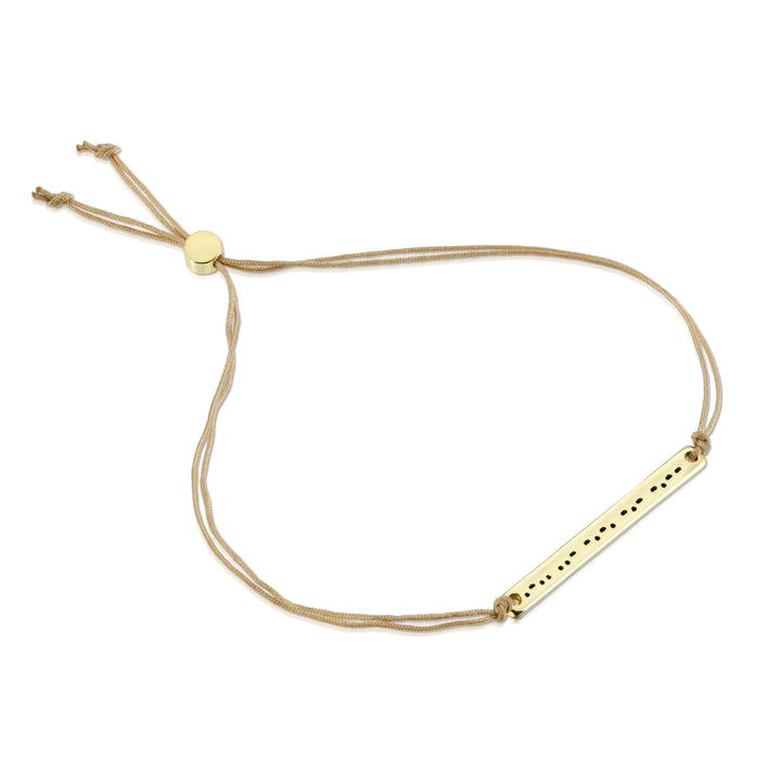 A gold-colored bracelet with a bar inscribed with dot-and-dash patterns on a beige cord.