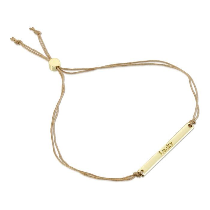 A gold bar bracelet with the word 'Lucky' inscribed on it hangs on a beige cord.