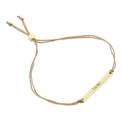 A gold bar bracelet with the word 'Lucky' inscribed on it hangs on a beige cord.