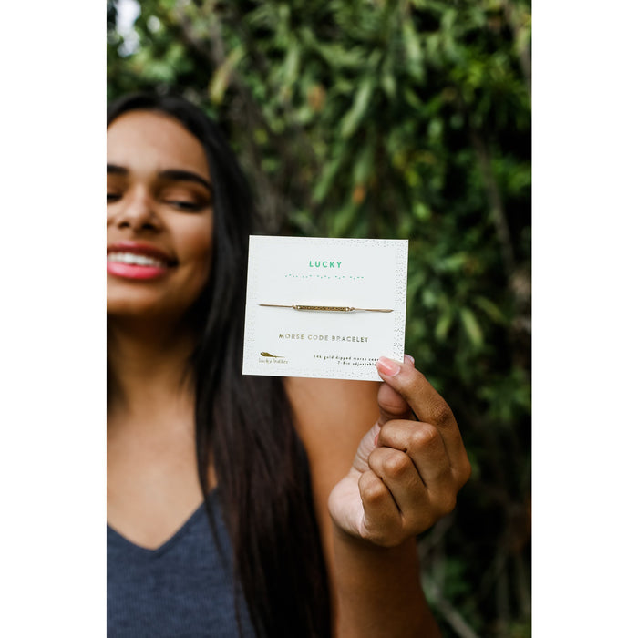 A woman out of focus holds a 'Lucky Feather' card for a 'Morse Code Bracelet' that says 'Lucky.'