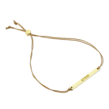 A gold-colored bracelet with the word 'QUEEN' engraved on it, attached to a light brown cord.