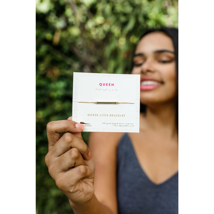 A person holds a product card for a 'Queen' morse code bracelet in front of green foliage.