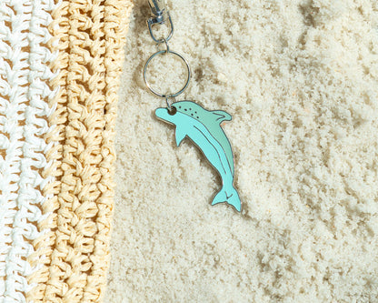 A straw bag with a turquoise dolphin keychain lies on sand.