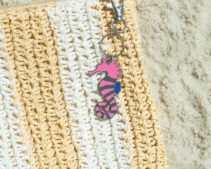 A woven straw bag with a pink seahorse keychain sits on sand.