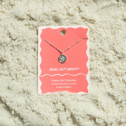 A 'Lucky Feather' necklace from the Ocean Life Collection is displayed on a pink card against a background of sand.