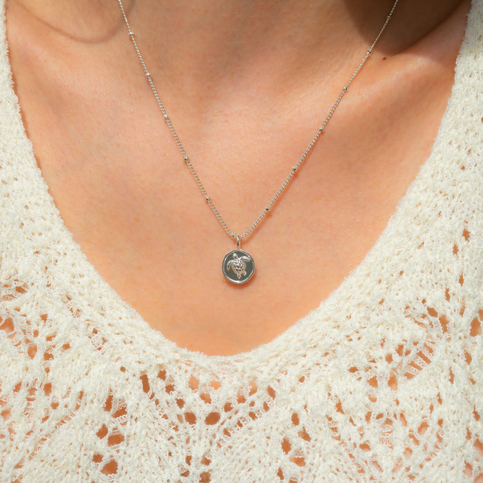 Close-up of a person wearing a silver chain necklace with a round pendant featuring a turtle.