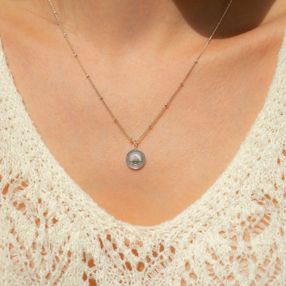 Close-up of a person's chest wearing a silver necklace with a seashell pendant and a lacy white top.