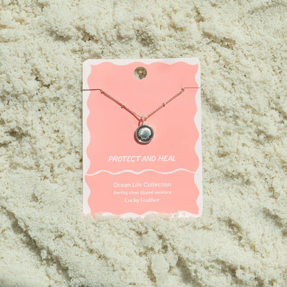 A seashell pendant necklace from Lucky Feather's Ocean Life Collection is displayed on a product card set on textured white sand.