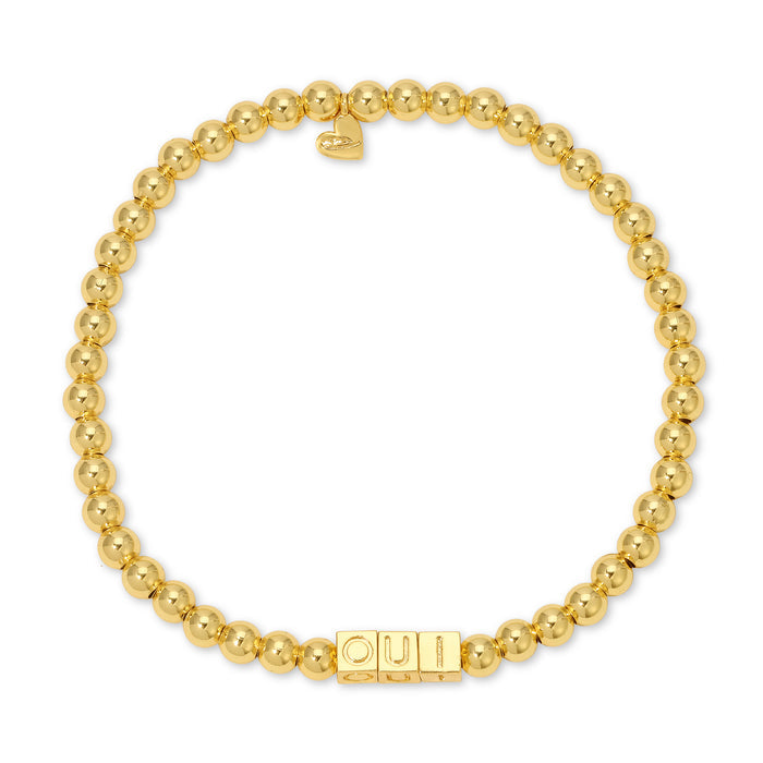 A gold-beaded stretch bracelet features the word 'OUI' spelled out in block letter beads and a small heart charm.