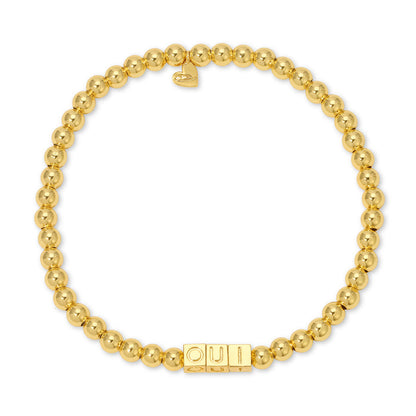 A gold-beaded stretch bracelet features the word 'OUI' spelled out in block letter beads and a small heart charm.