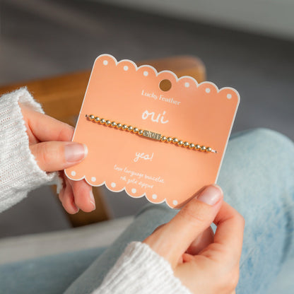 Hands hold a 'Lucky Feather' brand bracelet on a peach card with the word 'oui' on it.