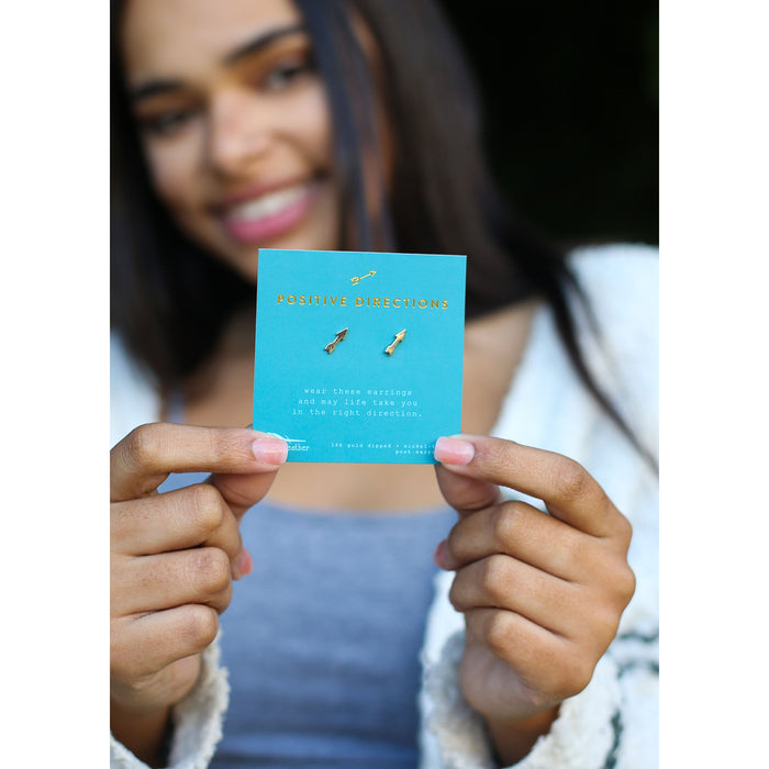 A smiling woman holds a product card for 'Positive Directions' earrings, which are gold-toned arrows.