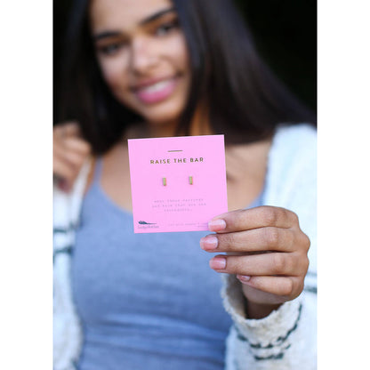 A smiling young woman holds a pink card with 'RAISE THE BAR' printed on it, displaying a pair of small gold earrings.