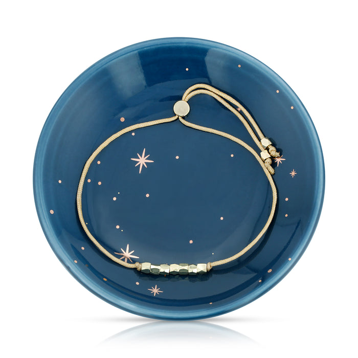 A gold bracelet with small, cubed, metallic beads rests on a blue, star-patterned dish.