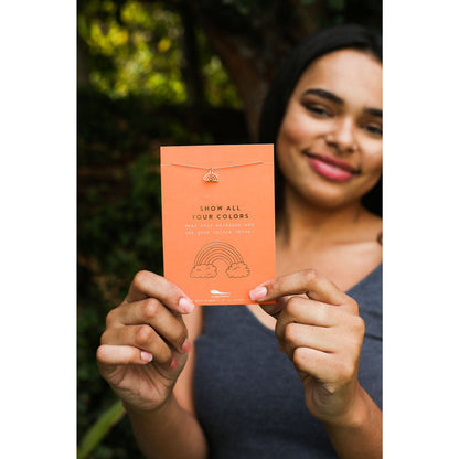 A woman smiles while holding up an orange card displaying a necklace with a rainbow charm.
