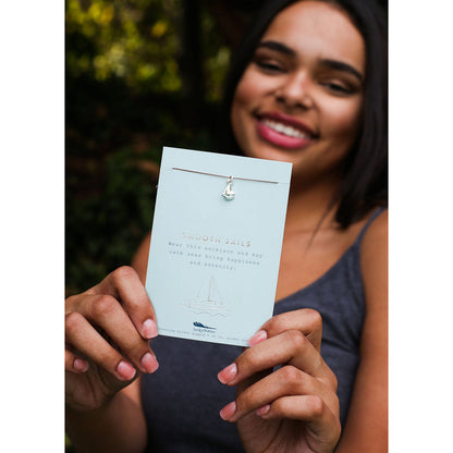 A smiling woman holds up a necklace with a sailboat charm on a card that reads 'Smooth Sails.'