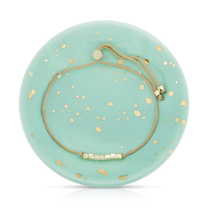 A gold-toned bracelet with geometric beads rests on a light turquoise ceramic plate with gold accents.