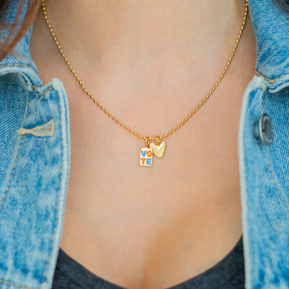 A person wears a gold chain necklace with a 'VOTE' charm and a gold heart.