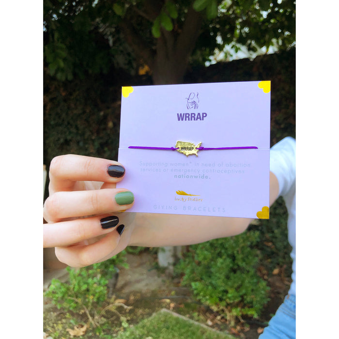 A hand holds a purple 'Lucky Feather' giving bracelet on a lavender card with the WRRAP logo.