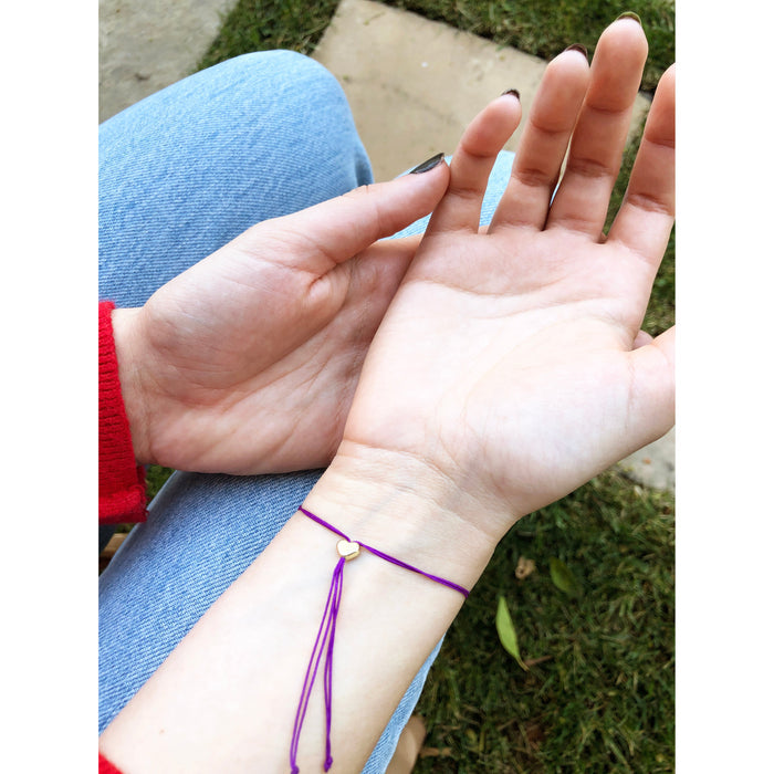 A person wearing a purple bracelet with a small gold heart charm is sitting in blue jeans.
