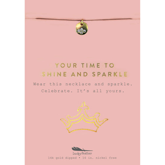 A necklace with a gold charm is on a pink display card with a crown graphic.