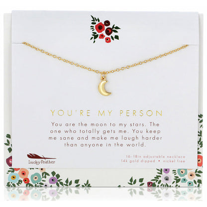 A gold-colored crescent moon pendant hangs from a thin chain displayed on a card with floral accents and the text 'You're my person.'