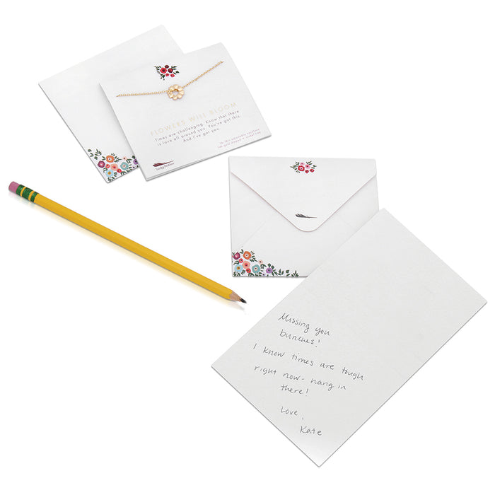A handwritten note with a yellow pencil, floral-themed stationery, and a necklace are displayed on a white surface.