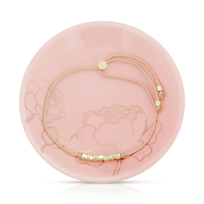 A delicate bracelet with gold beads on a light pink decorative plate with floral designs.